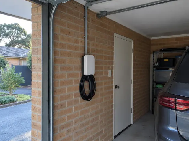 Wall-mounted EV charger with coiled cable on a brick garage wall beside a parked car in Kellyville