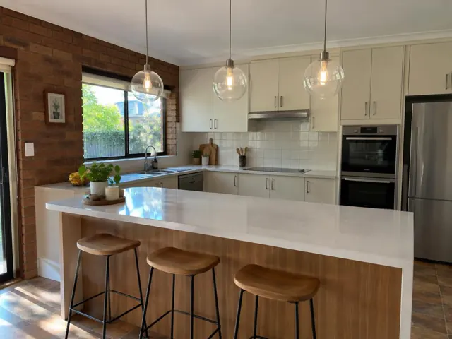 Kitchen with three glass pendant lights over a white island, brick feature wall and cream cabinetry in Cherrybrook